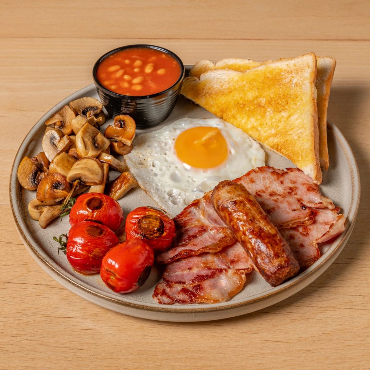 Full English breakfast: fried egg, bacon, sausage, grilled tomatoes, mushrooms, beans and toast on a stoneware plate