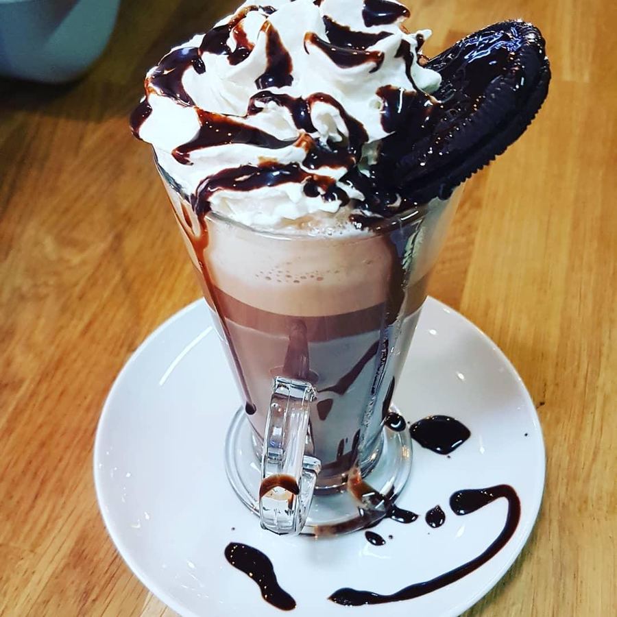 A latte glass of hot chocolate topped with whipped cream, chocolate sauce and an Oreo
