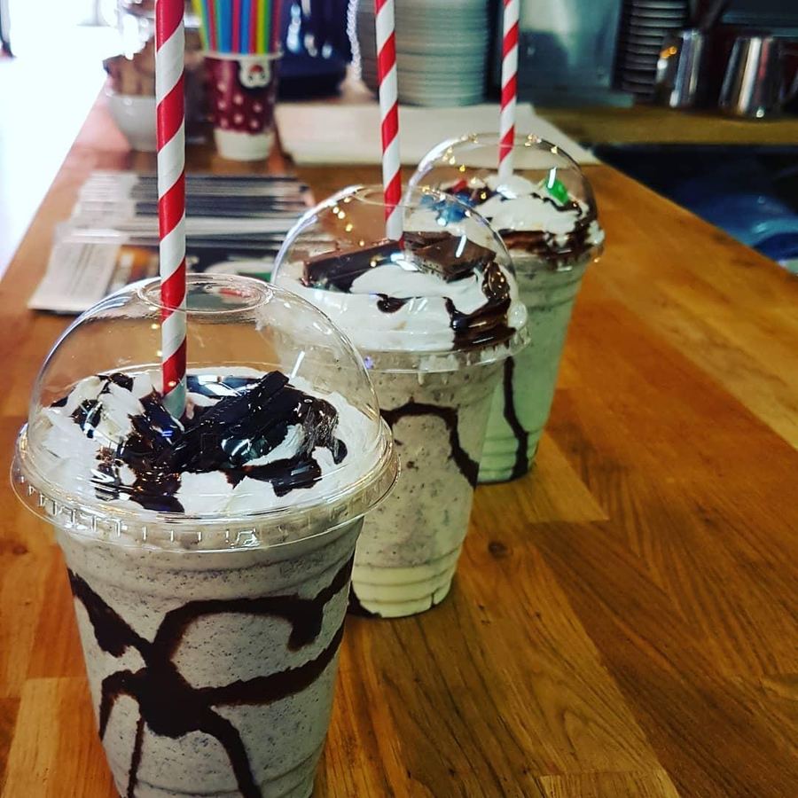Three tall milkshakes with cream, chocolate pieces and striped paper straws on the wooden counter